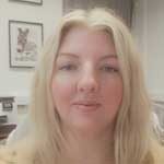 Blonde-haired woman looking at camera stood in front of wall with pictures hanging on it