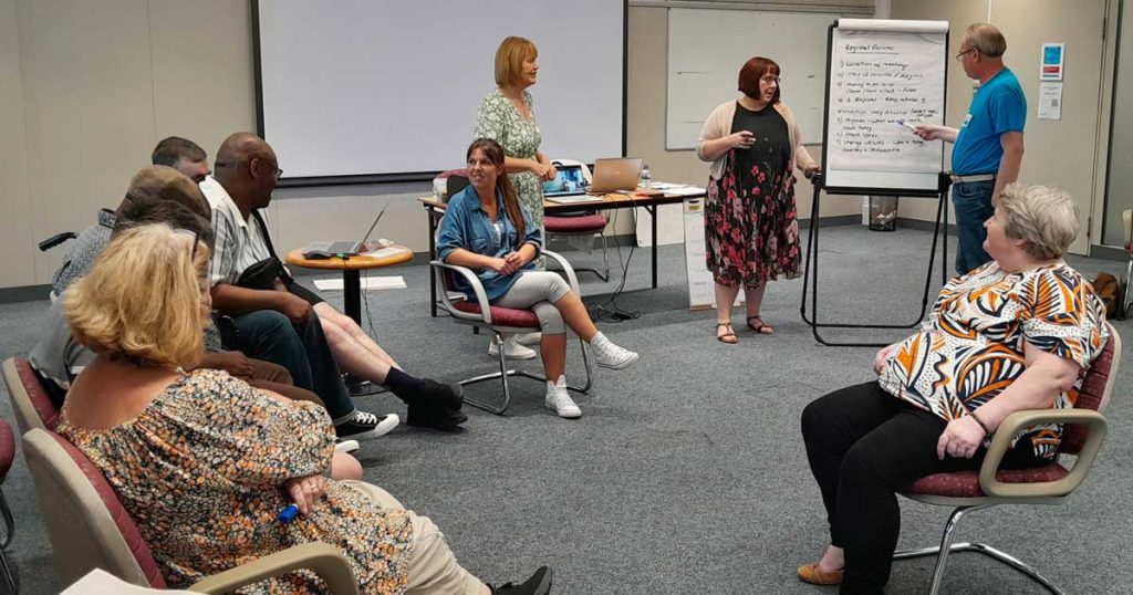 A man in a bright blue t-shirt indicates a sentence written on a flipchart