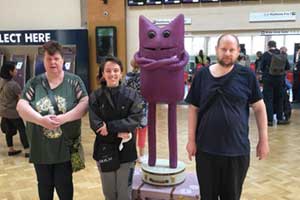 Three people wearing dark clothes stood next to a purple cat statue at a train station