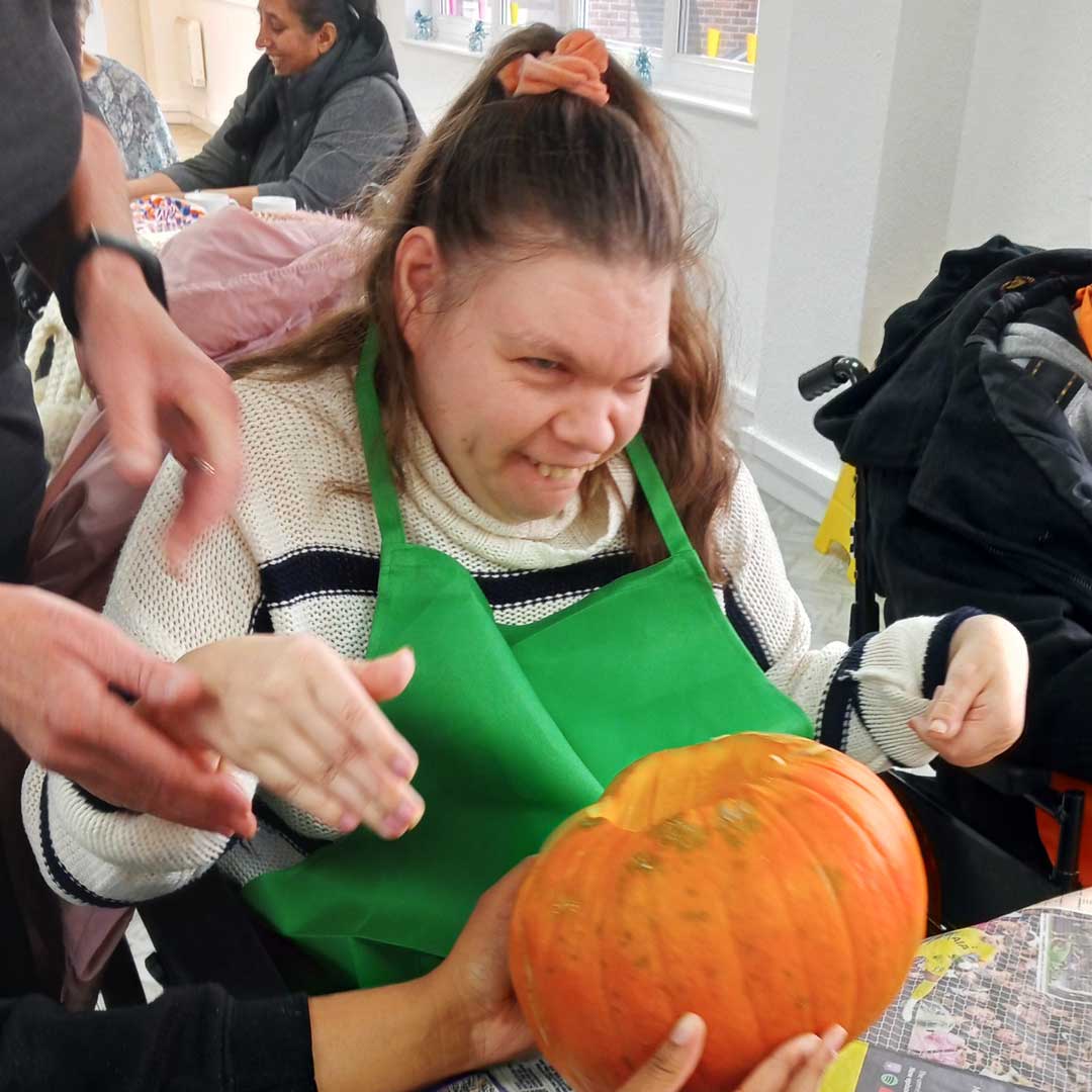 Woman wearing white jumper with a black stripe and a green apron about to pick up a pumpkin