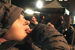 Woman wearing hooded black puffy jacket sat under floodlights at nighttime
