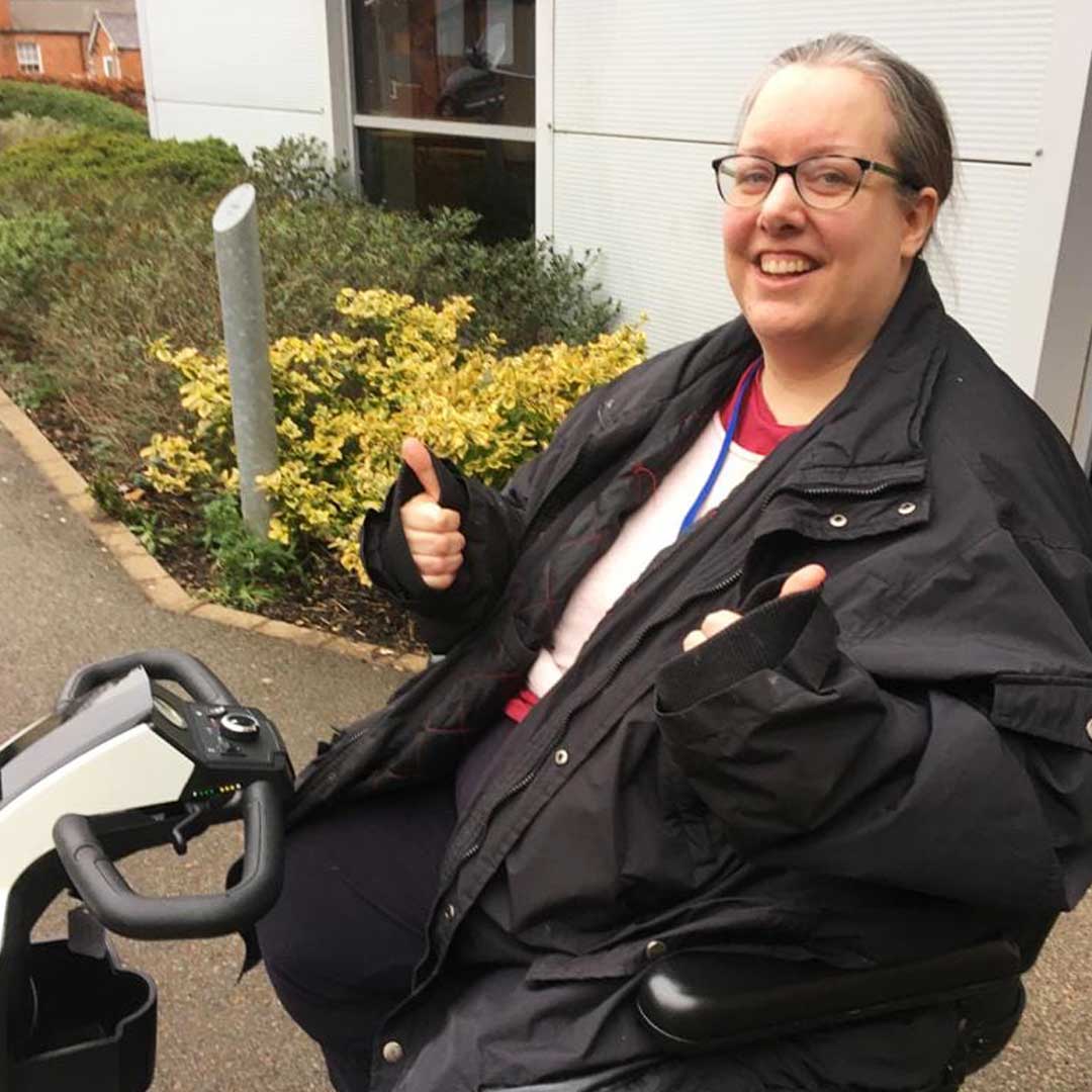 Woman wearing black jacket sat on a white mobility scooter outdoors giving the camera two thumbs up