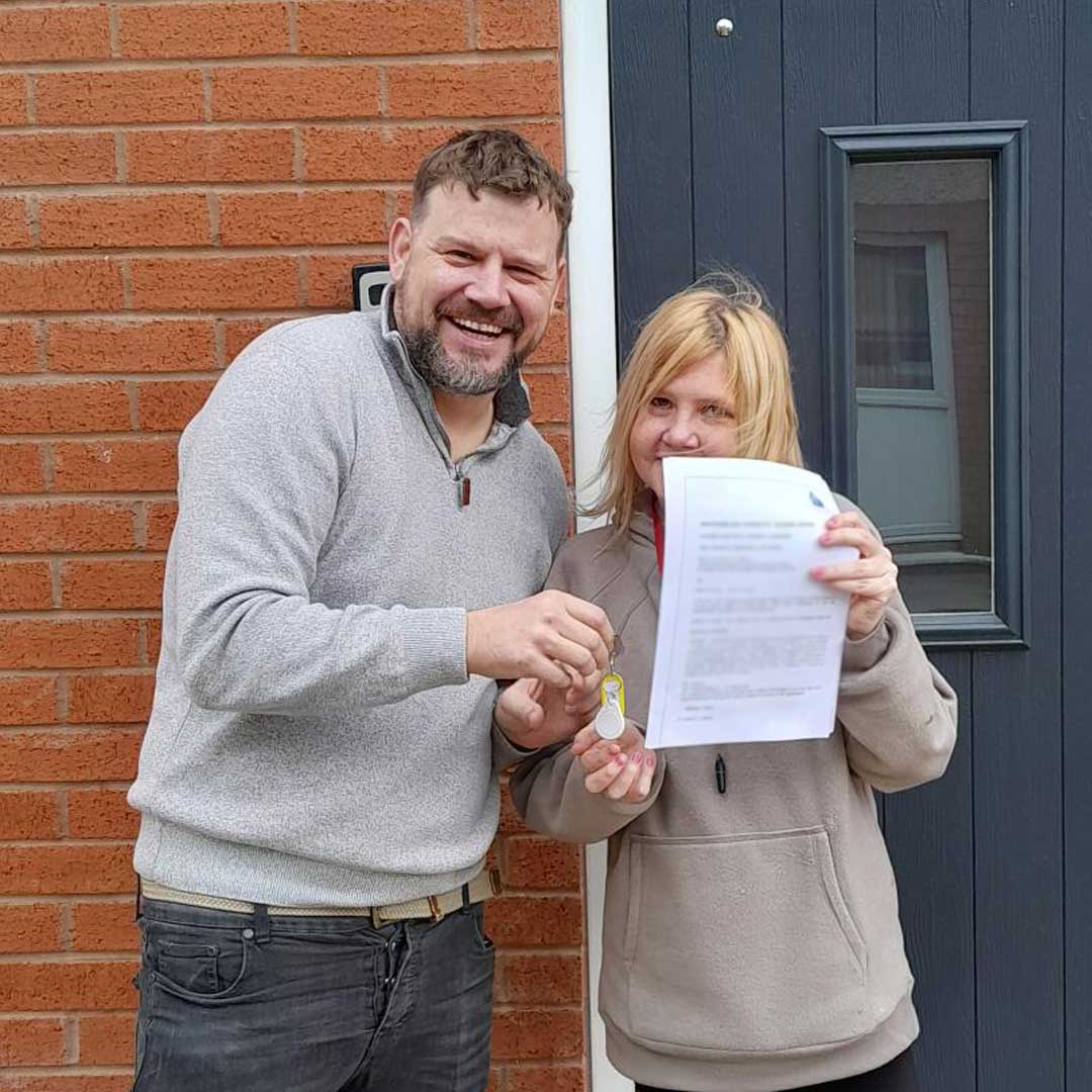 Man in grey jumper hands keys over to woman in beige jumper who's holding a tenancy agreement up to the camera