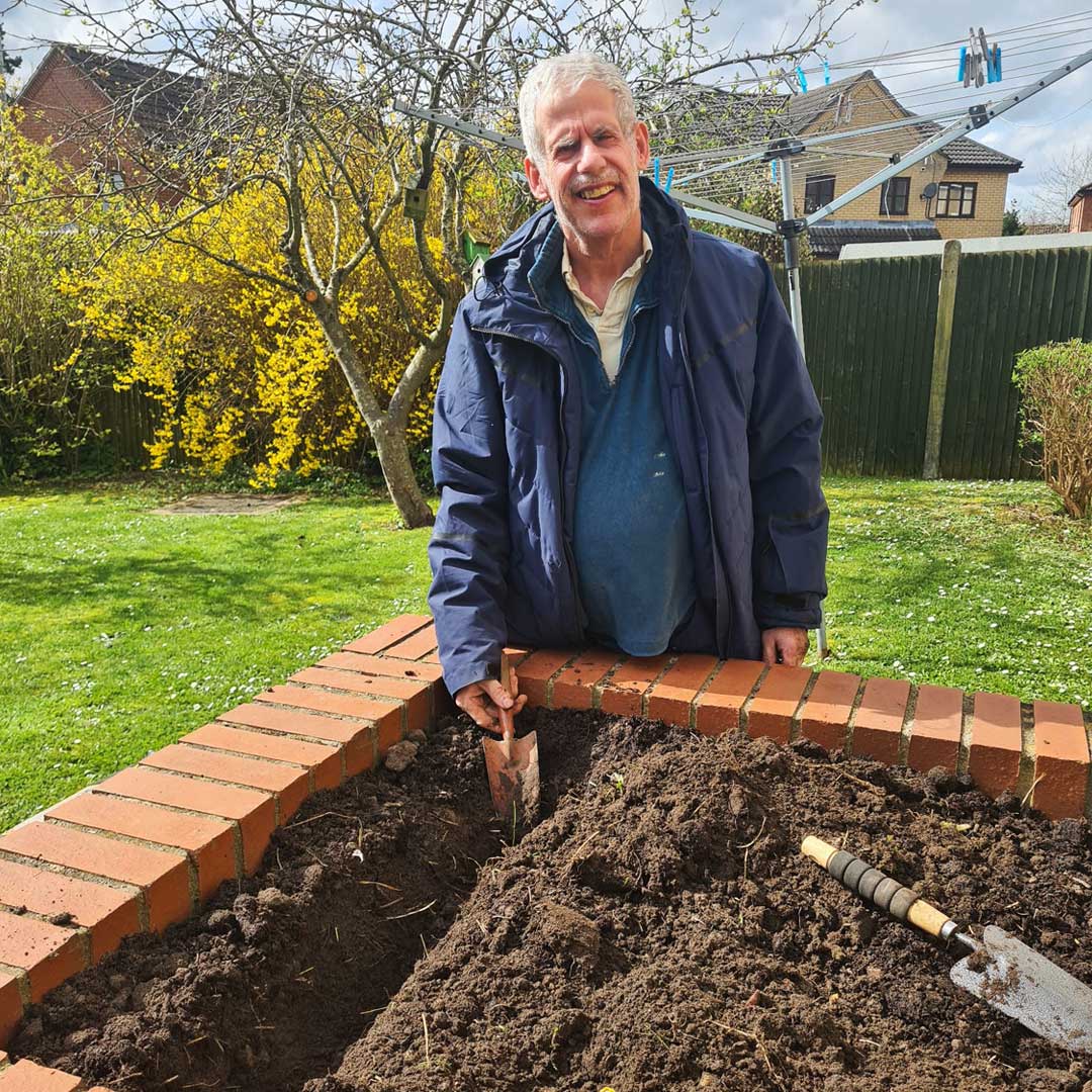 Man wearing navy blue jacket stood by a raised garden bed holding a trowel and smiling for the camera