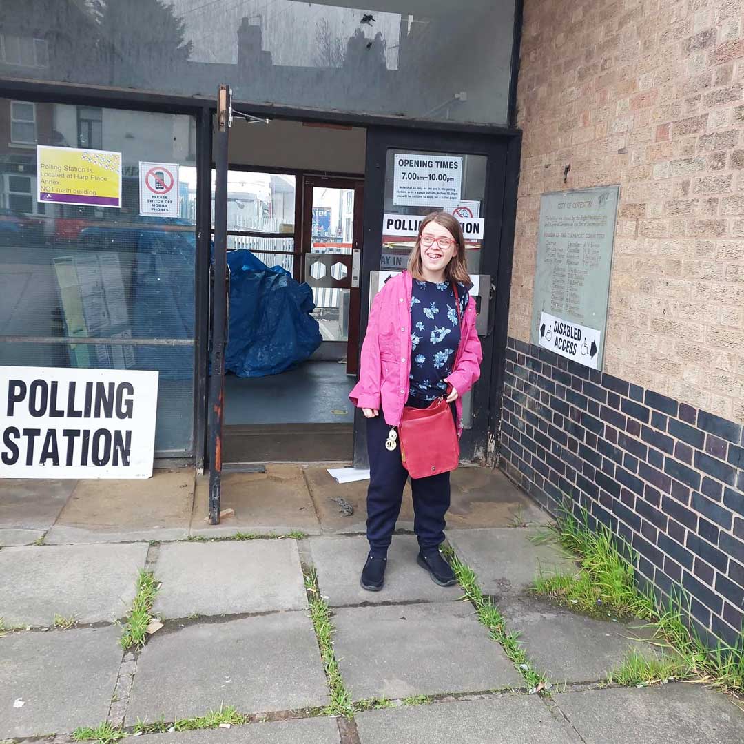 Woman wearing bright pink jacket and dark trousers stands outside a building next to a white sign with polling station written on it in black