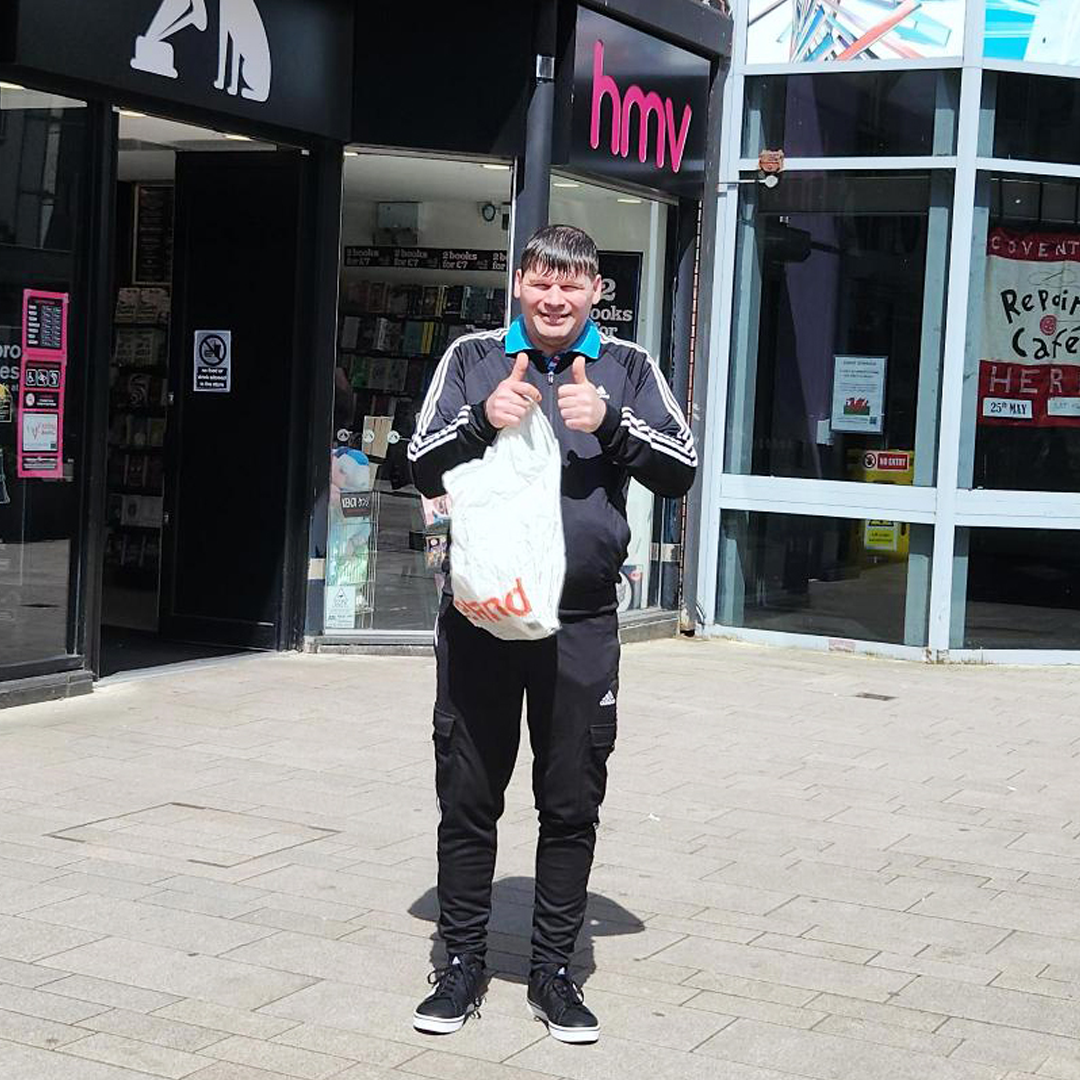 Man wearing black jacket with striped sleeves gives the camera a thumbs up, whilst stood in front of a shop holding a carrier bag