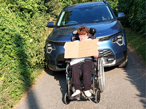 Woman in wheelchair holds up a cardboard placard in front of a car on a single track lane