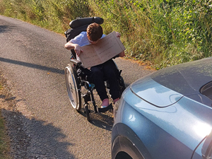 Woman in wheelchair holds up a cardboard placard in front of a car on a single track lane