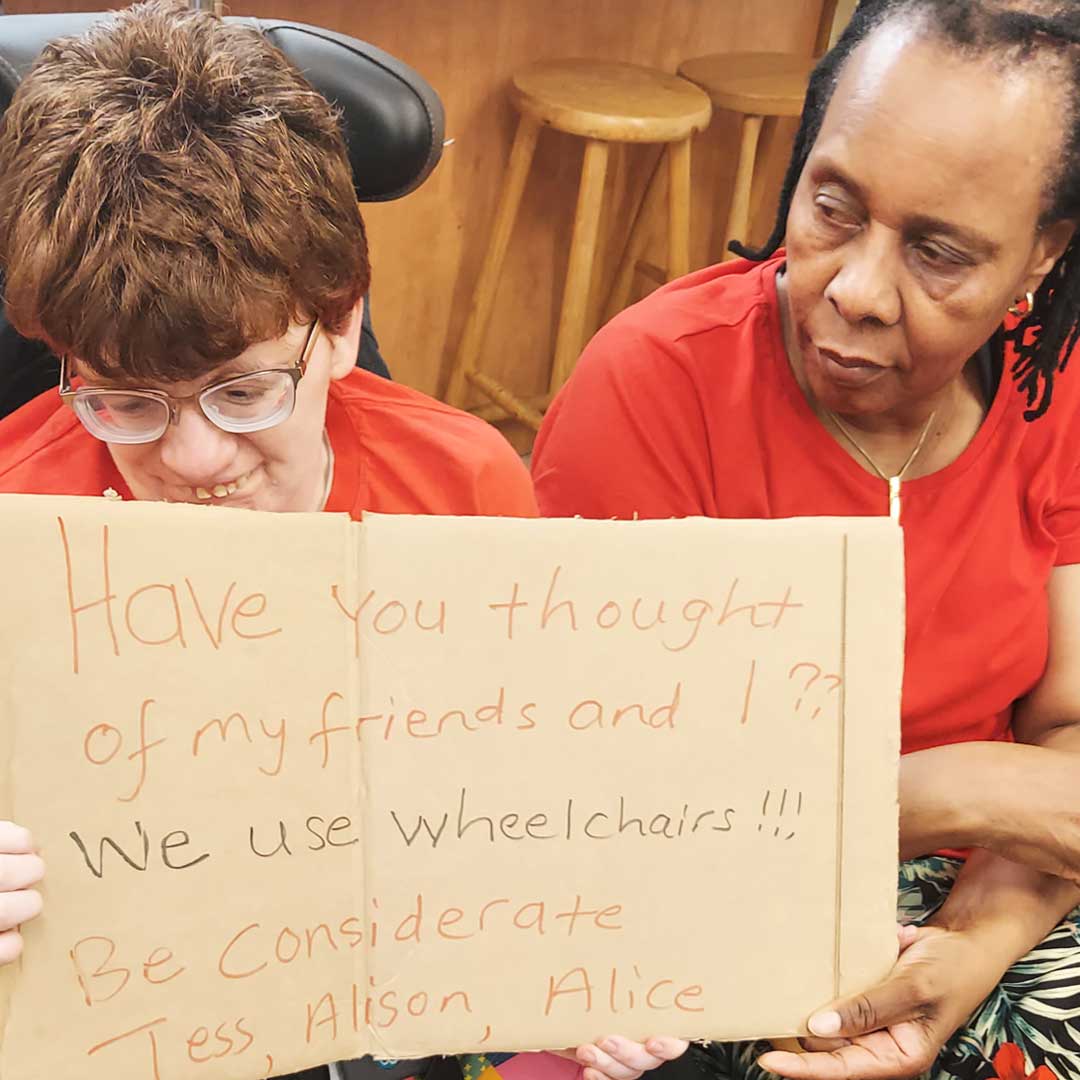 Woman in wheelchair wearing a red top holds up a cardboard placard, with her support worker sat next to her also wearing a red top