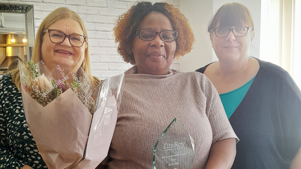 Three people smile for the camera, with one holding an award and one a bunch of flowers