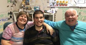 Joseph's mum and dad sat on a sofa with Joseph sat in between them. They are all grinning and his parents have their arms around his shoulders.