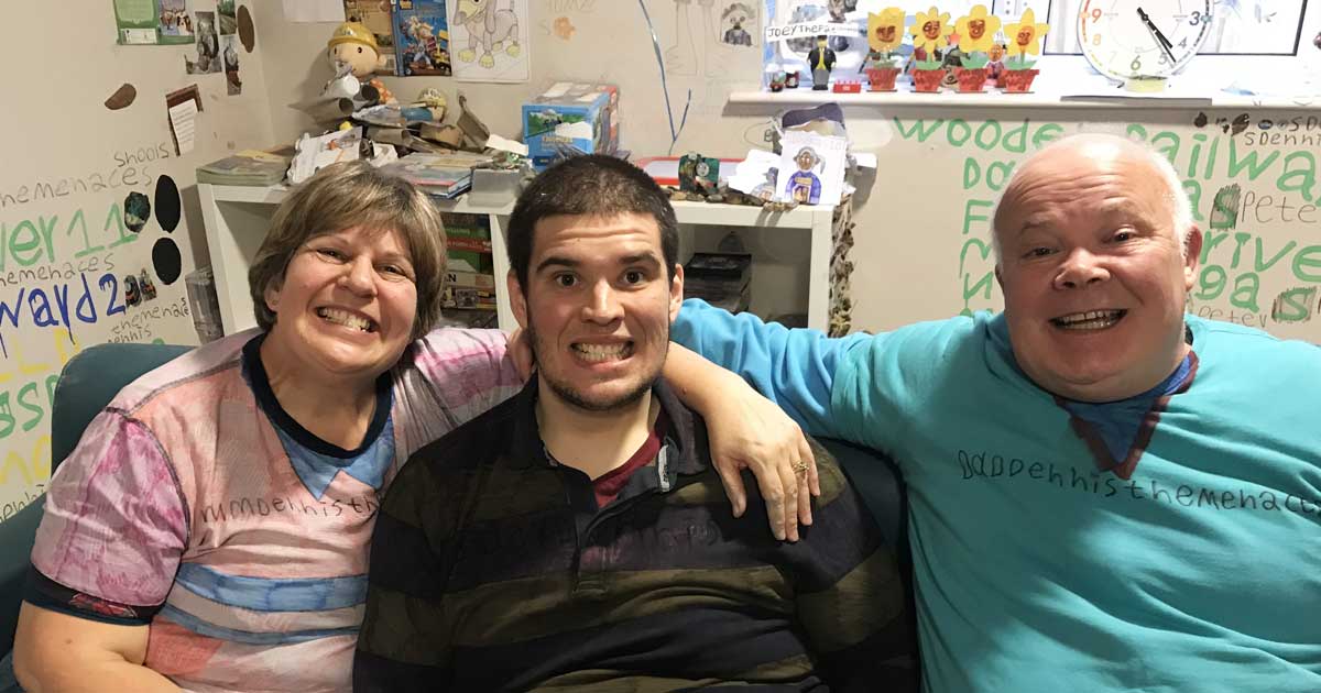 Two parents sat on a sofa with their adult son in between them, with their arms around his shoulders