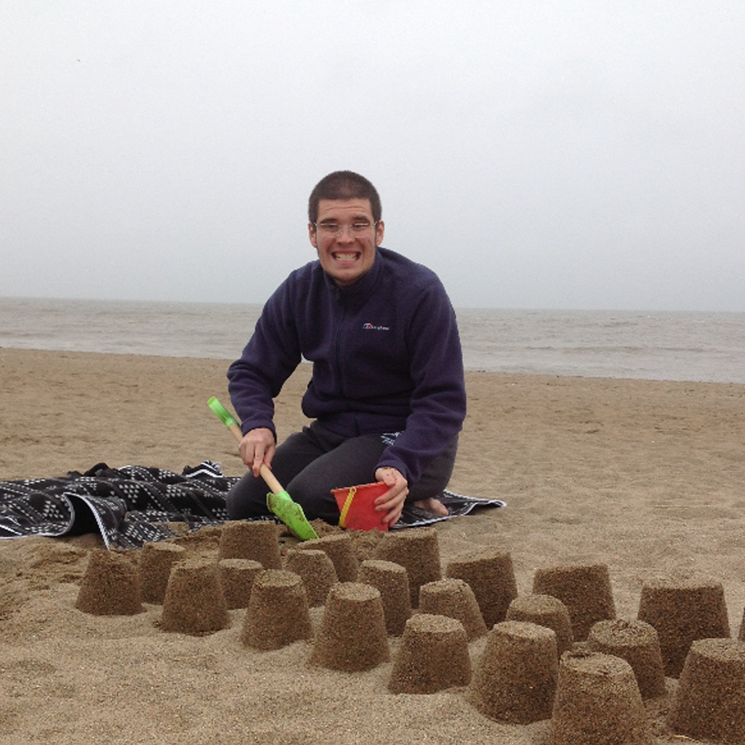 Young man on the beach wearing a dark sweater and smiling for the camera, with three lines of sandcastles in front of him