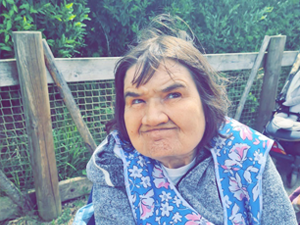 Woman wearing floral vest sat in front of a wooden and mesh fence with greenery behind it