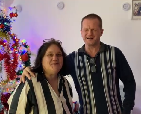 Couple stood in front of a white wall with a Christmas tree off to one side behind them