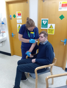 Lewis is pictured sitting in a chair. A woman in scrubs is holding his hand and holding a speculum near his mouth