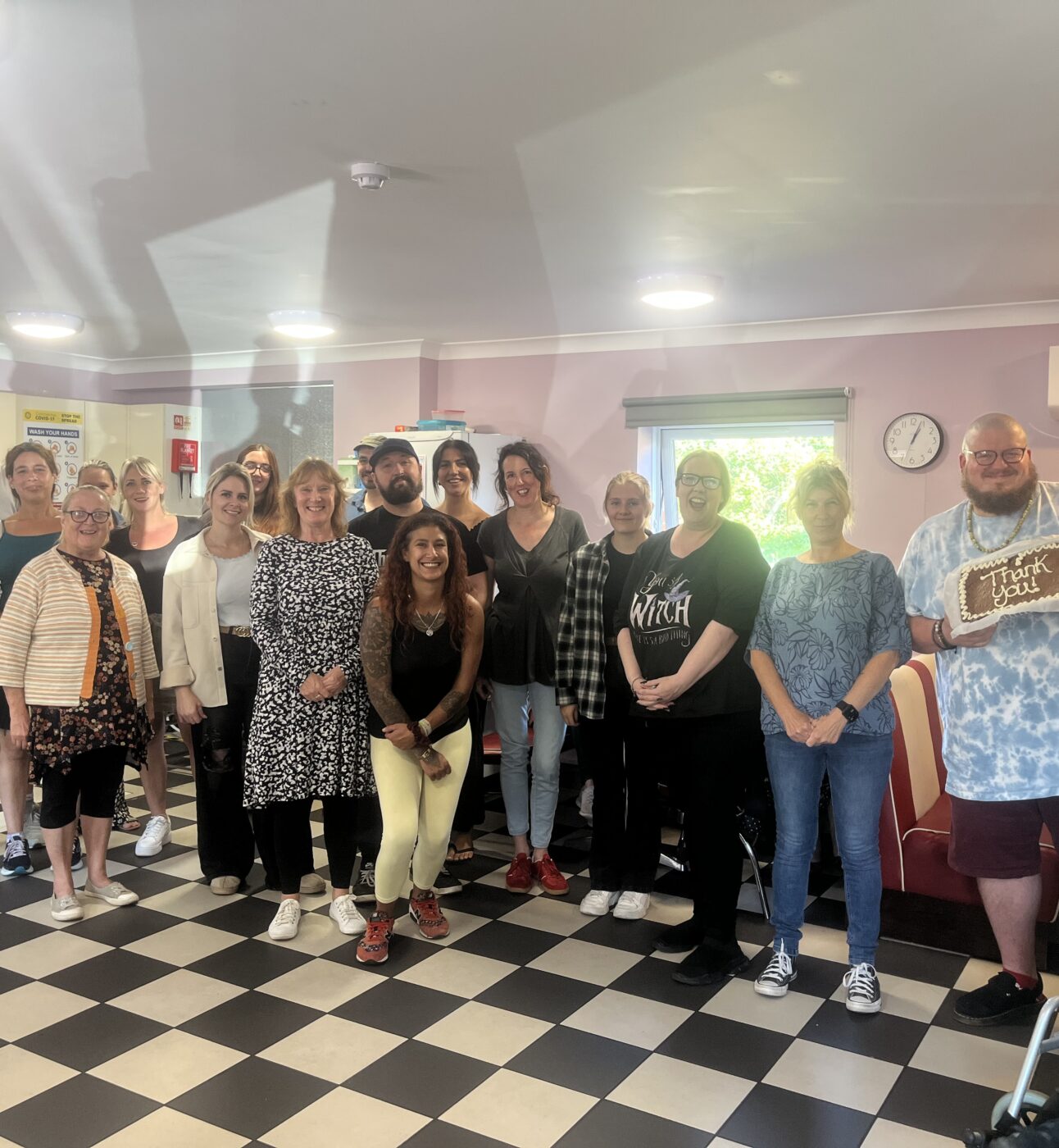 A large group photo of staff and members at The Maltings Day service, smiling and carrying two cakes with Thank You written on them