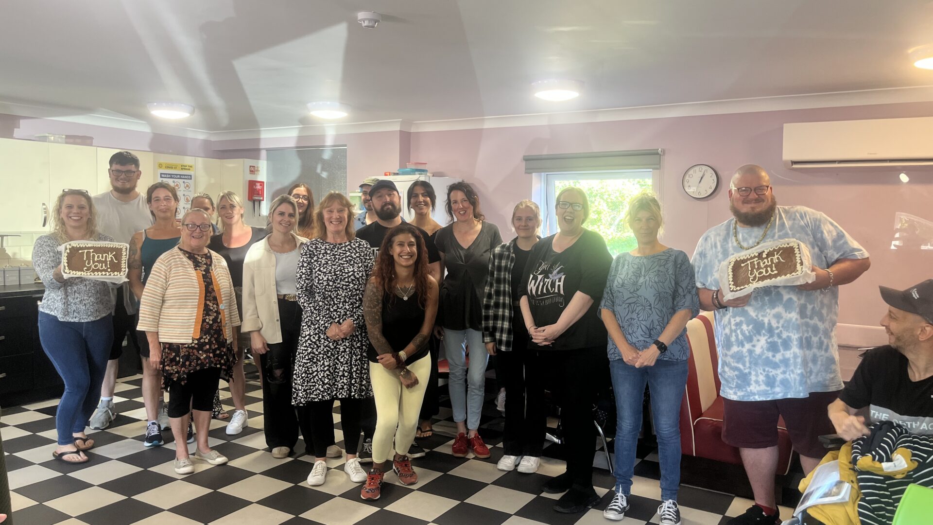 A large group photo of staff and members at The Maltings Day service, smiling and carrying two cakes with Thank You written on them
