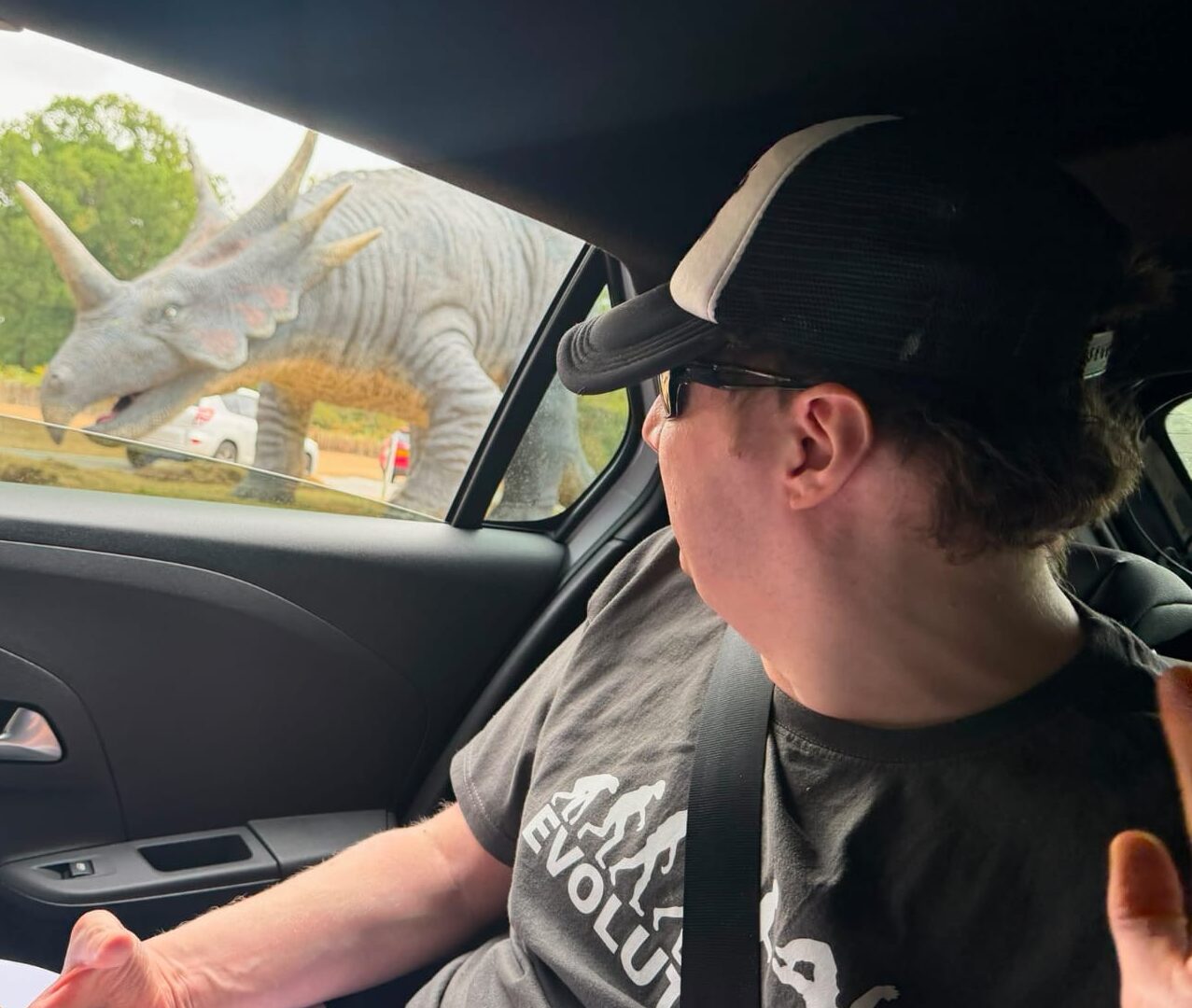 Alex is pictured in the back seat of a car. He is wearing a baseball cap and he is looking out the car window at a dinosaur.