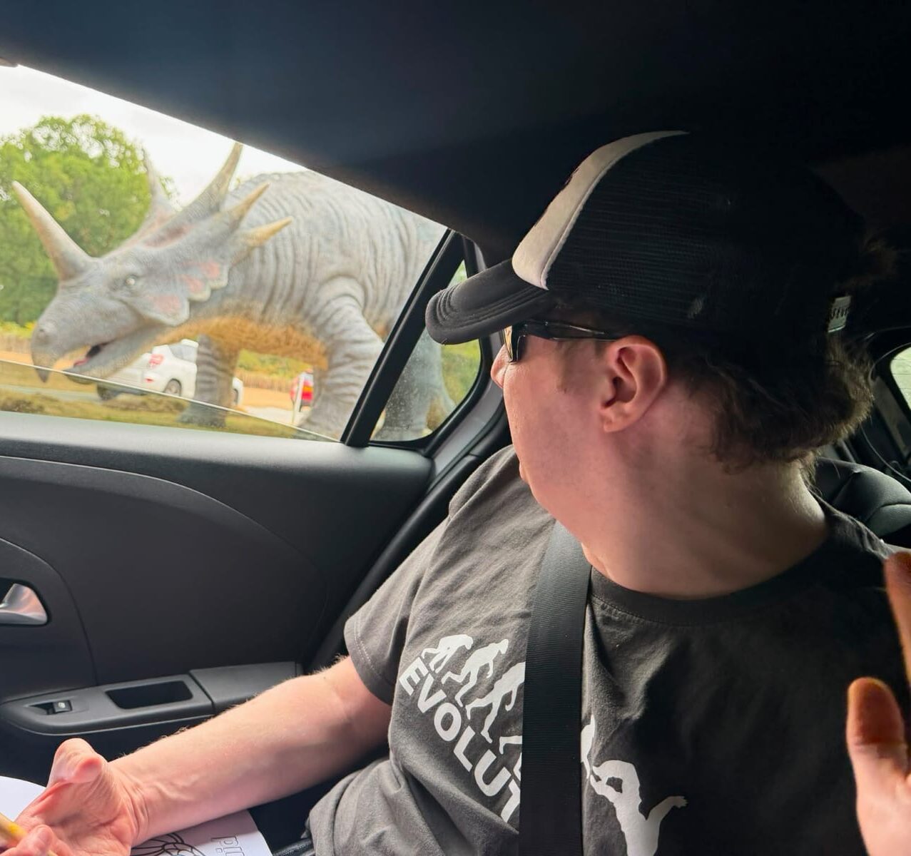 Alex is pictured in the back seat of a car. He is wearing a baseball cap and he is looking out the car window at a dinosaur.