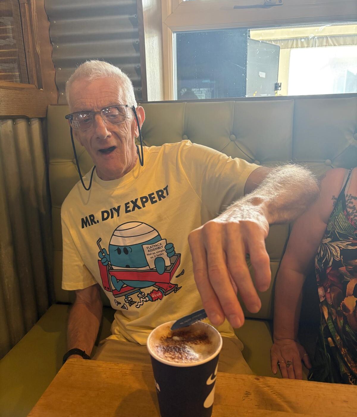 David is sitting in a cafe, putting sugar in his coffee with a smile on his face.