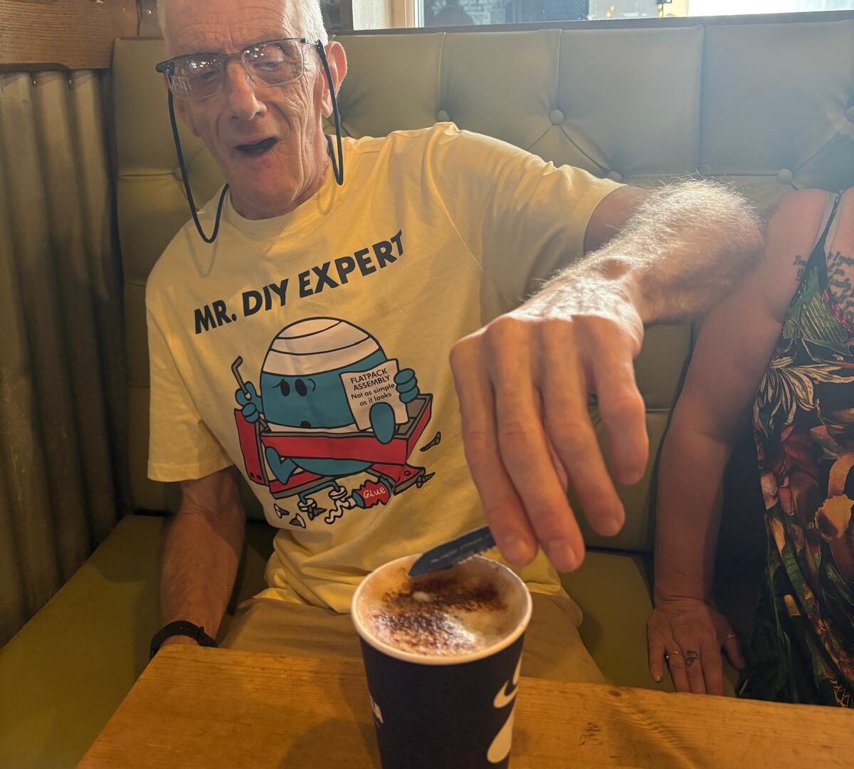 David is sitting in a cafe, putting sugar in his coffee with a smile on his face.