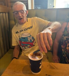 David is sitting in a cafe, putting sugar in his coffee with a smile on his face.